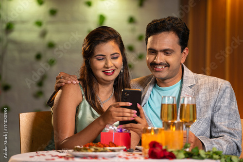 Happy Indian couples at candlelight dinner using mobile phone at restaurant - concept of social media sharing, online app or application and anniversary celebration.