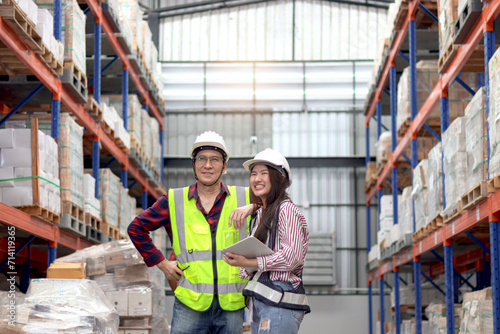 Happy Young Asian woman and senior man worker wearing safety vest and helmet, working at factory warehouse. Father and daughter work together in storage of family business.