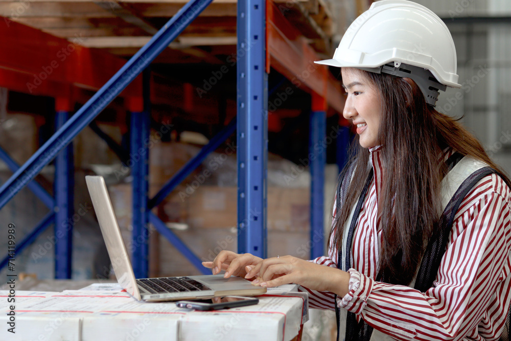 Side view of young Asian beautiful woman wearing safety vest and helmet, typing on a laptop computer at cargo logistics warehouse. female cargo logistics engineer working at storehouse workplace.