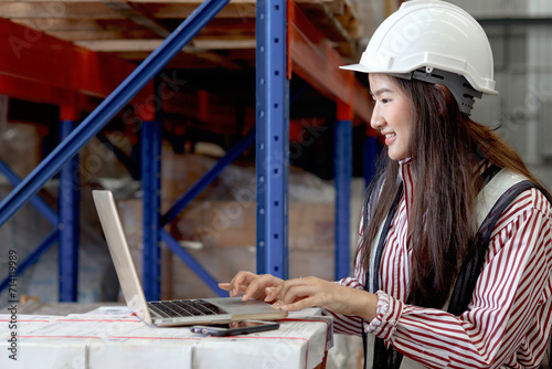 Side view of young Asian beautiful woman wearing safety vest and helmet, typing on a laptop computer at cargo logistics warehouse. female cargo logistics engineer working at storehouse workplace.