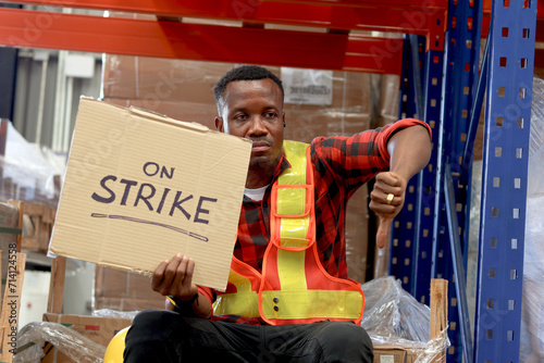 Angry unhappy African worker man wearing safety vest and giving thumb down with strike banner placard sign at cargo logistic warehouse. Striking worker protesting at workplace.