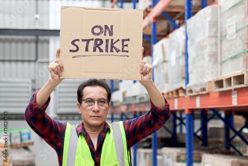 фотографія Angry unhappy Asian senior worker wearing safety vest, holding and raising sign on strike banner placard at cargo logistic warehouse