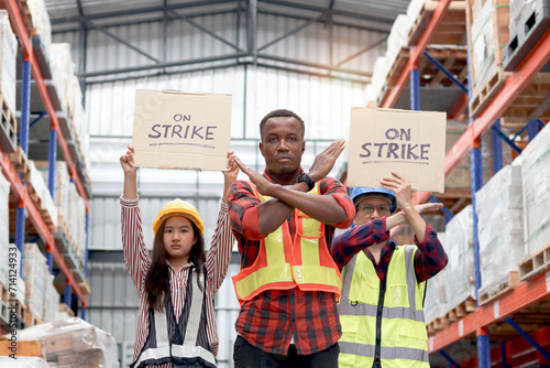 Angry unhappy African worker with colleagues, Asian senior and woman staffs wear safety vest and helmet, hold sign on strike banner at cargo logistic warehouse. Striking worker protesting at workplace photo