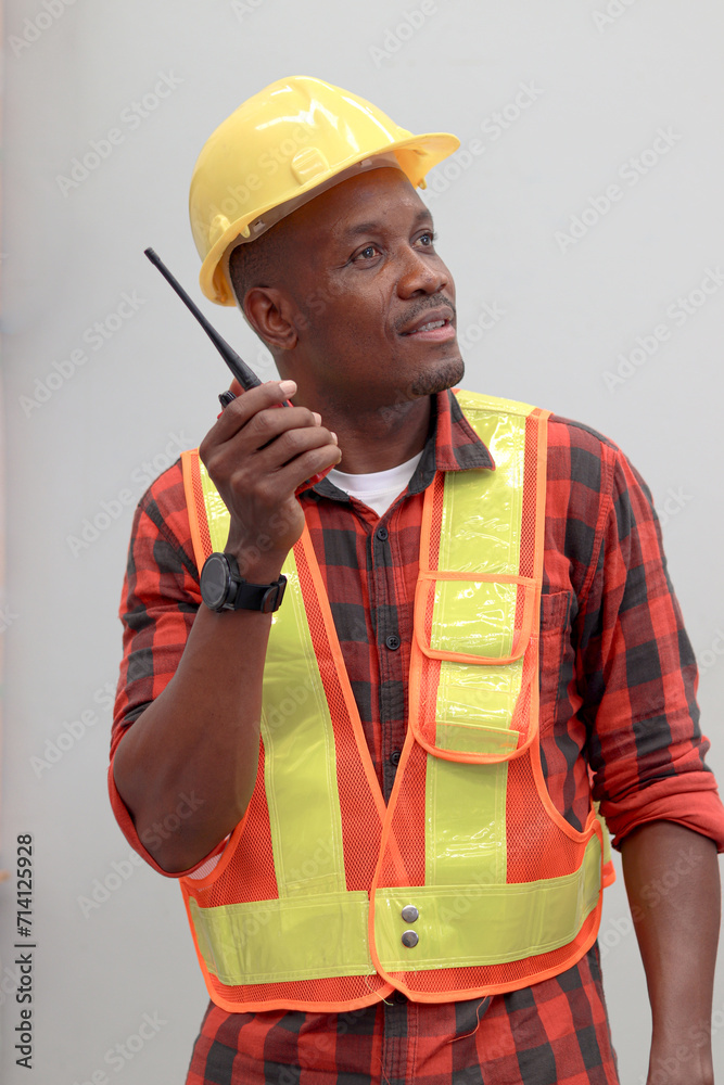 Portrait of young African worker man wearing safety vest and helmet, holding walkie talkie on gray walls. Engineer talking to a colleague with walkie talkie at logistic shipping cargo warehouse.