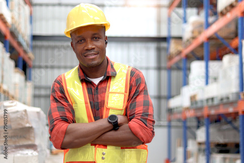 Portrait of young African worker man wearing safety vest and helmet, standing with arms crossed at warehouse factory. Male cargo logistics staff working at shipping storehouse workplace.