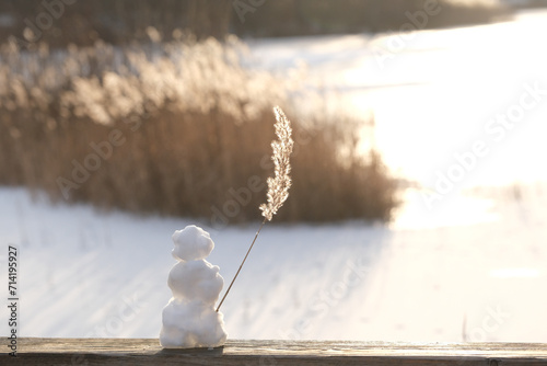 Little Snowman standing on bridge in front of a frozen lake waving with a reed in backlight of sinking winter sun like a seaman on his ship waving goodbye to maybe the last snow.