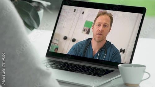 A handsome male doctor in blue scrubs consults a virtual patient over laptop, he discusses symptoms and then prescribes medicine. 