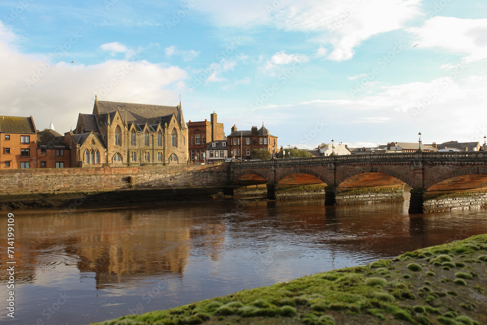 An ancient stone bridge over a river in a Scottish town with historical buildings and a Gothic church in the background.