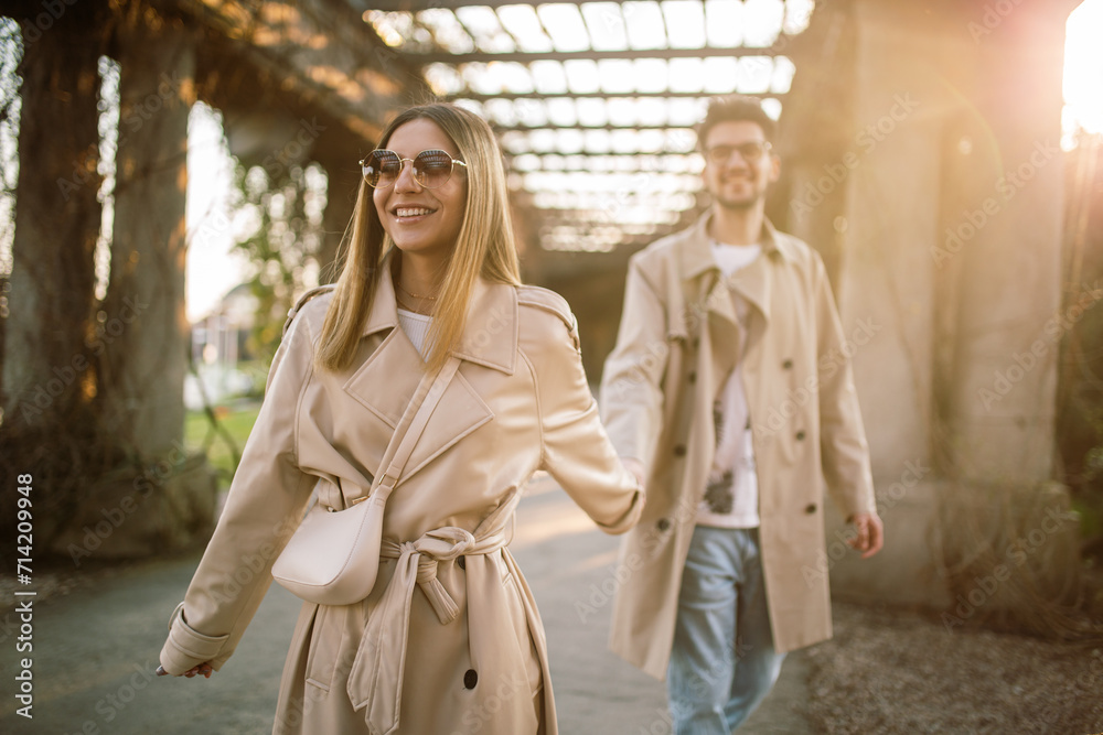 Young couple having fun in the park at sunset on a sunny autumn warm day. Stylish man and woman in beige trench coats.