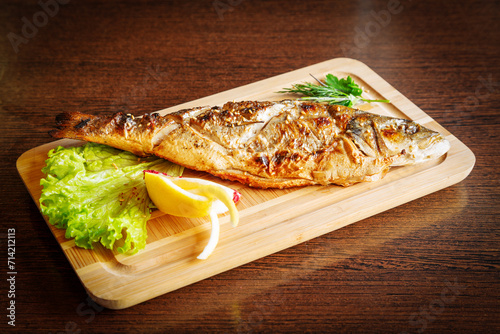 Whole grilled fish with lemon and lettuce on a wooden board.