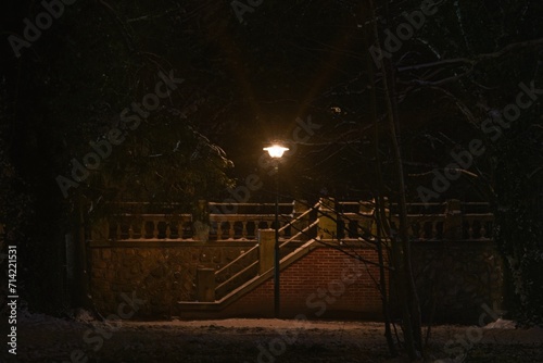 Zimowy wieczór w parku. Samotna, elektryczna lampa oświetlająca kamienne schody o konstrukcji murowanej z czerwonej cegły. Ziemię pokrywa warstwa białego śniegu.