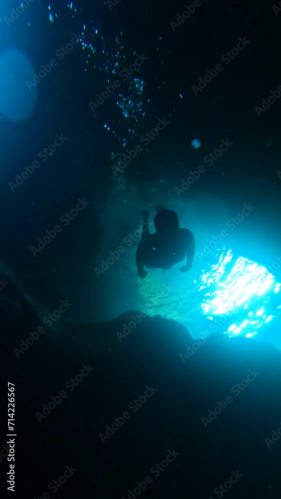 Explore the depths of an underwater cave in slow motion. A young diver, with goggles, glides towards the cave's depths, with sunbeams piercing through its two openings, and air bubbles ascending. Explore the depths of an underwater cave in slow motion. A young diver, with goggles, glides towards the cave's depths, with sunbeams piercing through its two openings, and air bubbles ascending.
