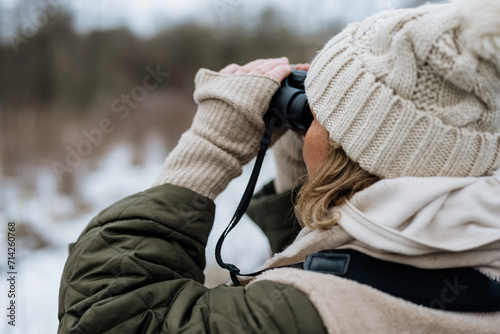 Woman dressed in winter clothes with gloves and knitted beanie looking for birds and animals through the binoculars in nature photo