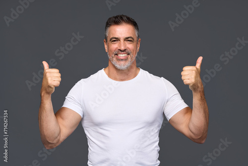 Happy fit sporty older man coach, middle aged personal trainer wearing white t-shirt showing like thumbs up standing isolated on gray background motivating for good results giving recommendation.