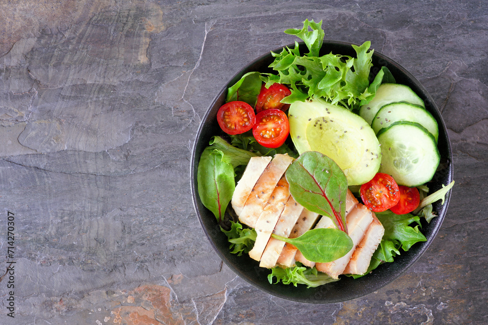 Healthy homemade salad bowl with avocado and chicken. Above view on a dark slate stone background.