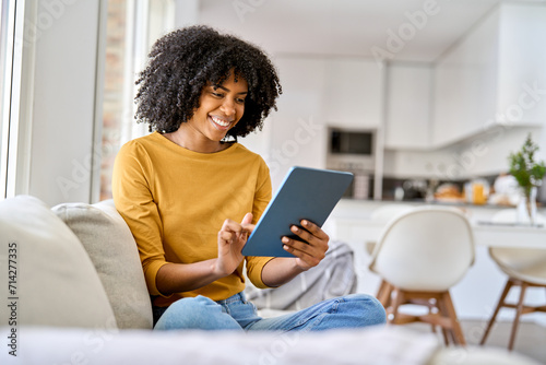 Happy young African American woman sitting on couch at home using digital tablet. Smiling relaxed lady relaxing on sofa looking at tab technology device holding pad computer in hands in living room.