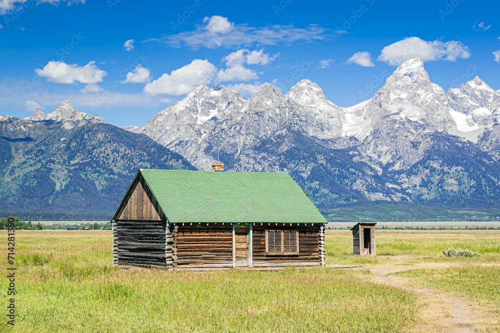 Historic wood log building in the valley meadow in Grand Teton National Park with the snow-capped Teton Mountain Range and blue sky with clouds in the background.