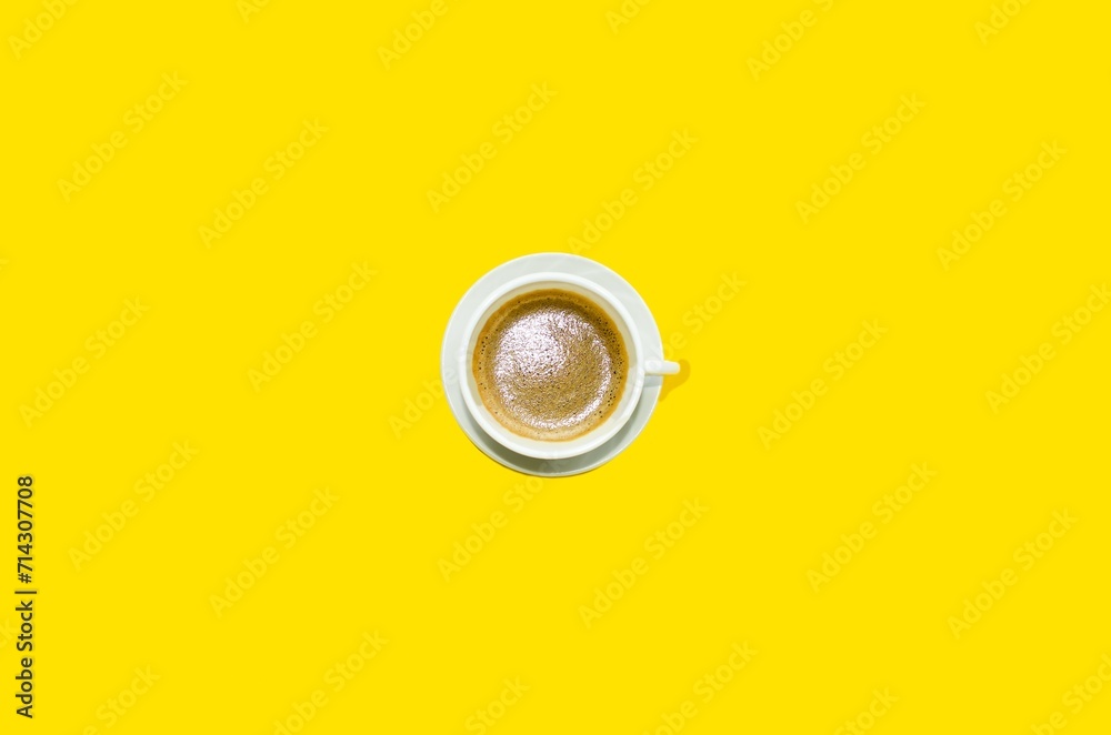On a bright yellow background in the center there is a white cup with black coffee, cappuccino.  Space for copy text, flat lay.  Fragrant, delicious hot coffee.  Top view, close-up.