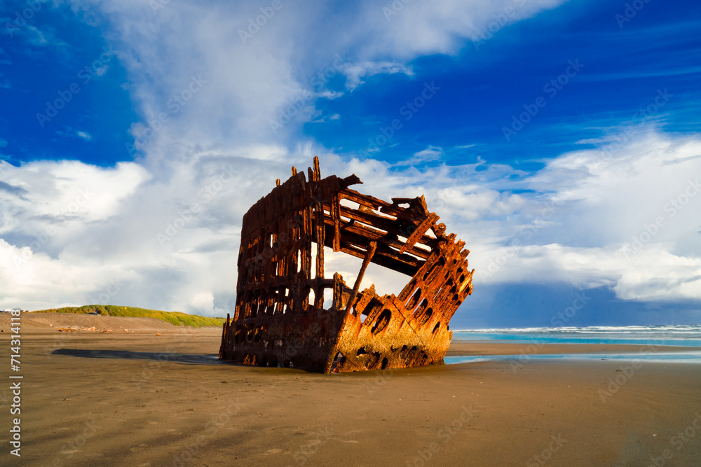 The skeleton of a shipwreck sits in the sand on the coast of Oregon on  a sunny day with rain clouds in the background. 