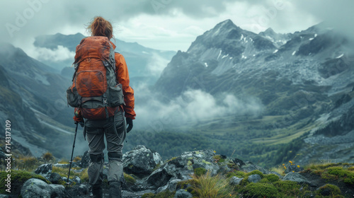  a person with a backpack standing on top of a mountain looking at a valley with snow capped mountains in the distance with low clouds in the sky and low lying clouds.
