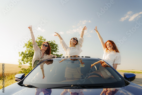Three friends pop out of a car sunroof, arms raised in joy, embracing the freedom of a summer road trip in the countryside © arthurhidden