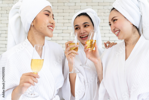 Three young women wearing bathrobes holding wine glasses stand talking in a bedroom