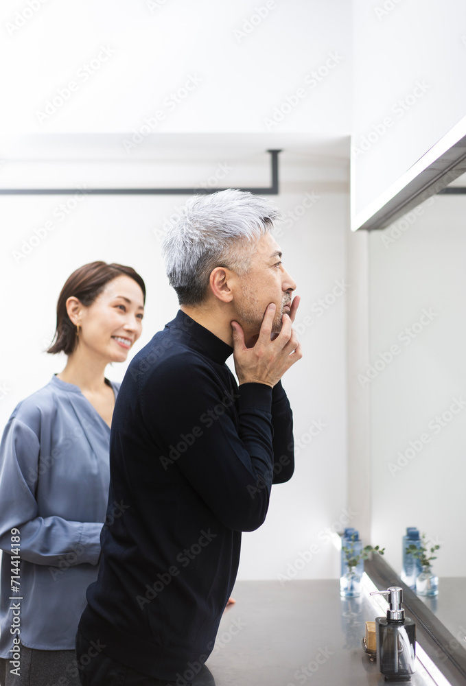鏡を見ながら顔を触っているミドル男性/女性がスキンケアを教えている/メンズビューティー 鏡を見ながら顔を触っているミドル男性/女性がスキンケアを教えている/メンズビューティー