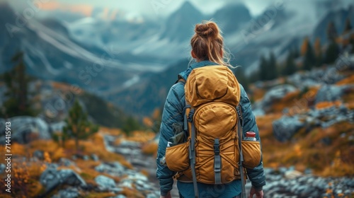  a woman with a backpack is walking up a mountain trail towards a valley with a mountain range in the distance and snow capped peaks in the distance in the distance.