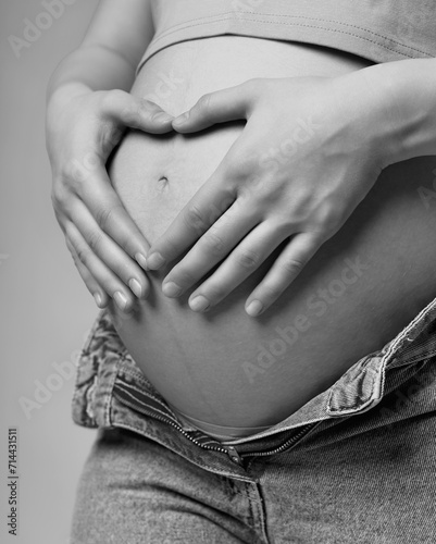 Black and white photo of female on 6th month of pregnancy. Woman touching with hands her naked belly and shows heart sign.