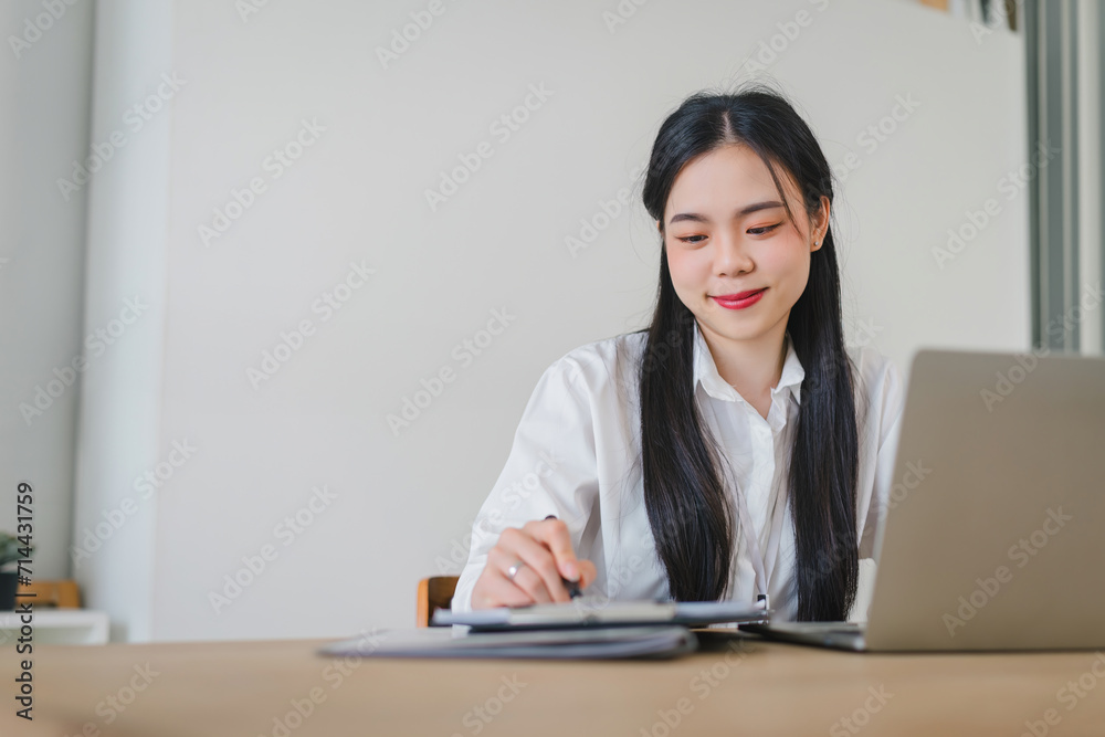 Focused asian professional businesswoman calculating budget using laptop and taking notes in a bright office environment. Focused asian professional businesswoman calculating budget using laptop and taking notes in a bright office environment.