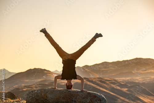 Active young man is standing upside down on big rock in sunset mountains photo