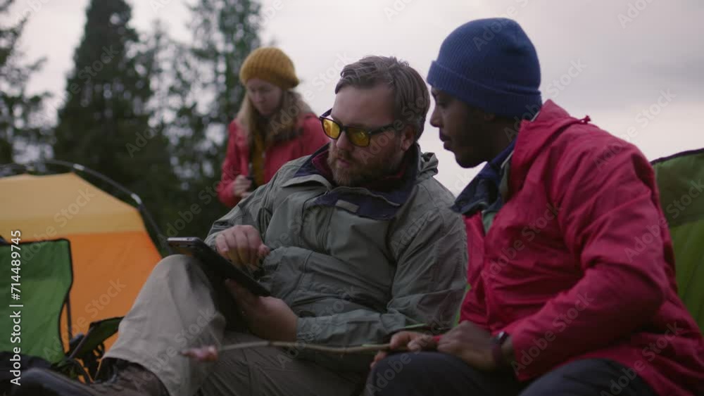 Female hiker packing up and prepares poles to next hike. Multiethnic men plan and discuss route. Caucasian tourist with African American friend checks trail on digital tablet computer. Active leisure. Female hiker packing up and prepares poles to next hike. Multiethnic men plan and discuss route. Caucasian tourist with African American friend checks trail on digital tablet computer. Active leisure.