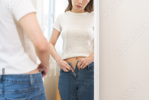 Excess weight gain, sad overweight young woman wearing fit trousers standing in front of mirror at home, hand trying to close zip up and button of jeans from fat around waist. Obese problem concept. photo