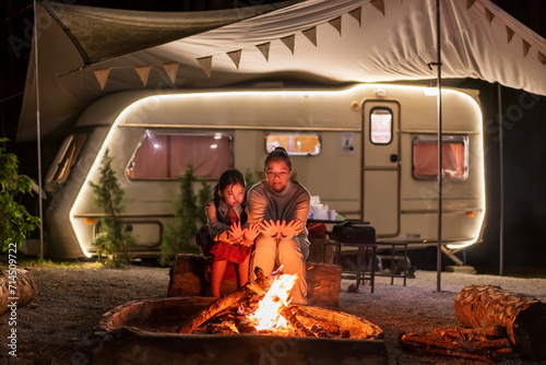 Asian family mother and child camper or kid girl travel camping sitting together on log to warming themselves by bonfire at night with campervan or RV motorhome in holiday at Doi Bo Luang Forest Park photo