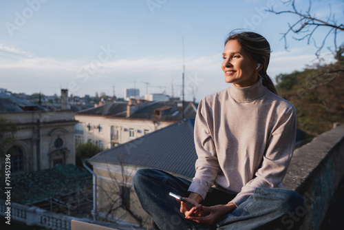 Woman freelancer uses laptop on cement wall outdoors against the sky and the roof of the city. The woman to be focused on her work or enjoying some leisure time while using her laptop. © svetograph