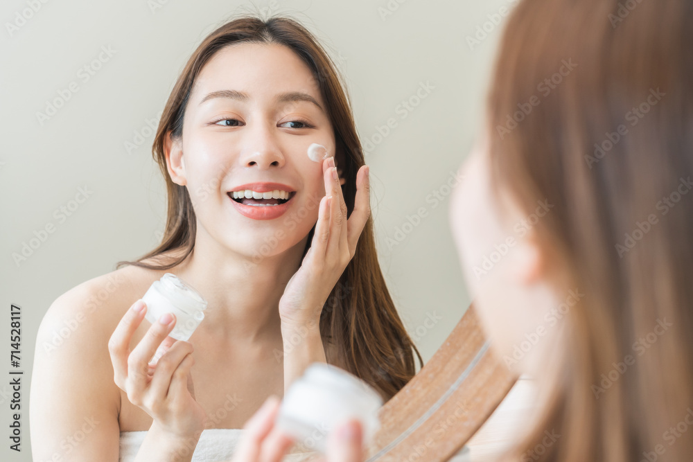 Facial beauty skin care, smile of pretty asian young woman in bathrobe looking at mirror, hand applying moisturizer lotion on her face, holding jar of skin cream before makeup cosmetic routine at home Facial beauty skin care, smile of pretty asian young woman in bathrobe looking at mirror, hand applying moisturizer lotion on her face, holding jar of skin cream before makeup cosmetic routine at home