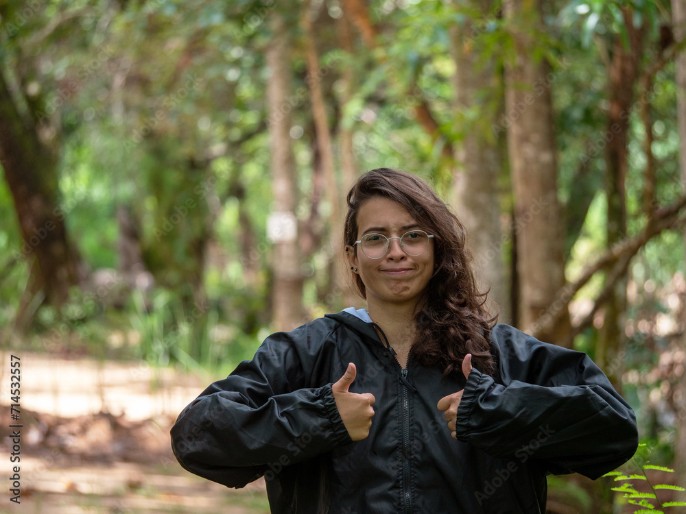 Jeune femme portant une veste imperméable se forçant à sourire avec les pouces en l'air dans une forêt Jeune femme portant une veste imperméable se forçant à sourire avec les pouces en l'air dans une forêt