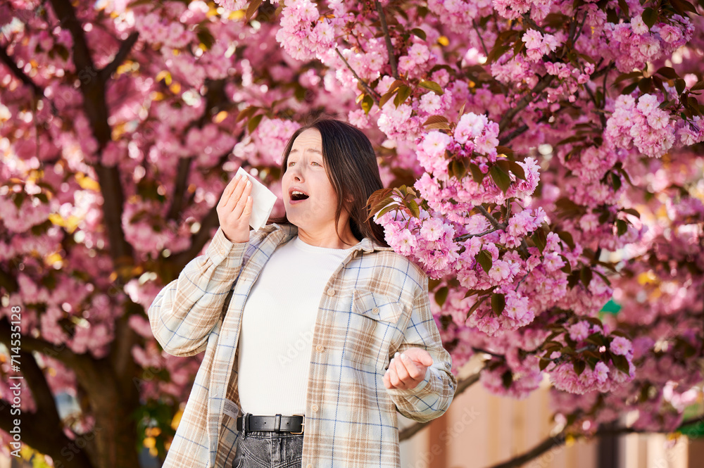 Woman allergic suffering from seasonal allergy at spring in blossoming garden at springtime. Woman sneezing and blowing nose using nasal handkerchief in front of blooming tree. Spring allergy concept. Woman allergic suffering from seasonal allergy at spring in blossoming garden at springtime. Woman sneezing and blowing nose using nasal handkerchief in front of blooming tree. Spring allergy concept.