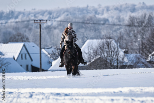 A young red-haired woman rides her horse at a walk across a Schebeckte meadow with a snowy village in the background on a sunny cold winter day.