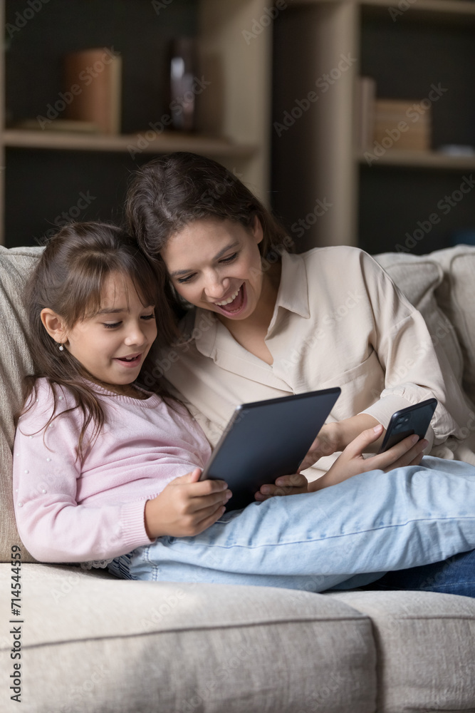 Vertical shot pretty woman and daughter sit on sofa at home with digital tablet gadget, spend free time on internet, buying via electronic commerce marketplace app, enjoy funny videos and application Vertical shot pretty woman and daughter sit on sofa at home with digital tablet gadget, spend free time on internet, buying via electronic commerce marketplace app, enjoy funny videos and application