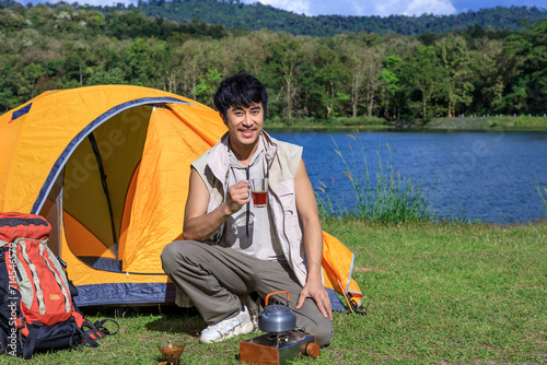Asian male man drinking coffee and feeling relax with camping tent beside lake and mountain, looking at camera