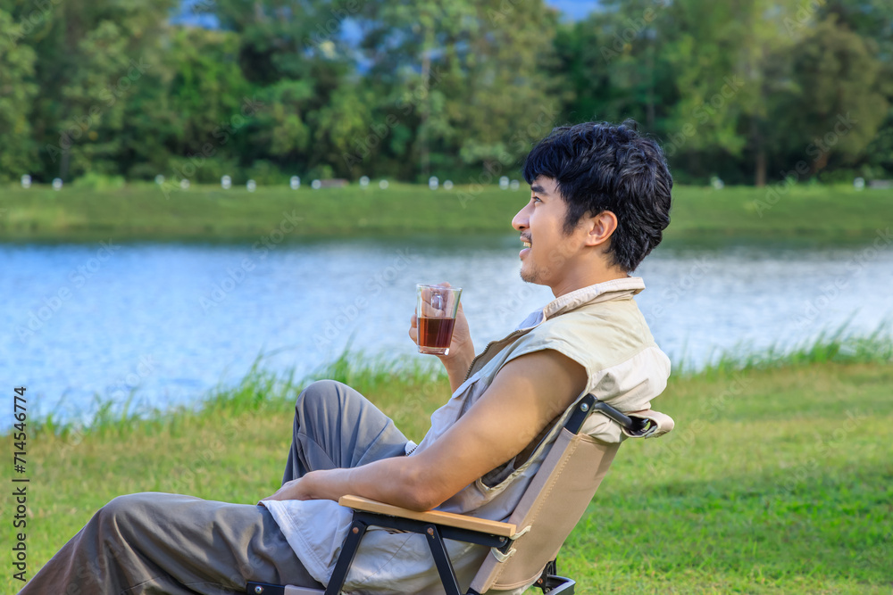 Fototapeta premium Asian male man sitting on chair, drinking coffee and feeling relax during camping beside lake and mountain.