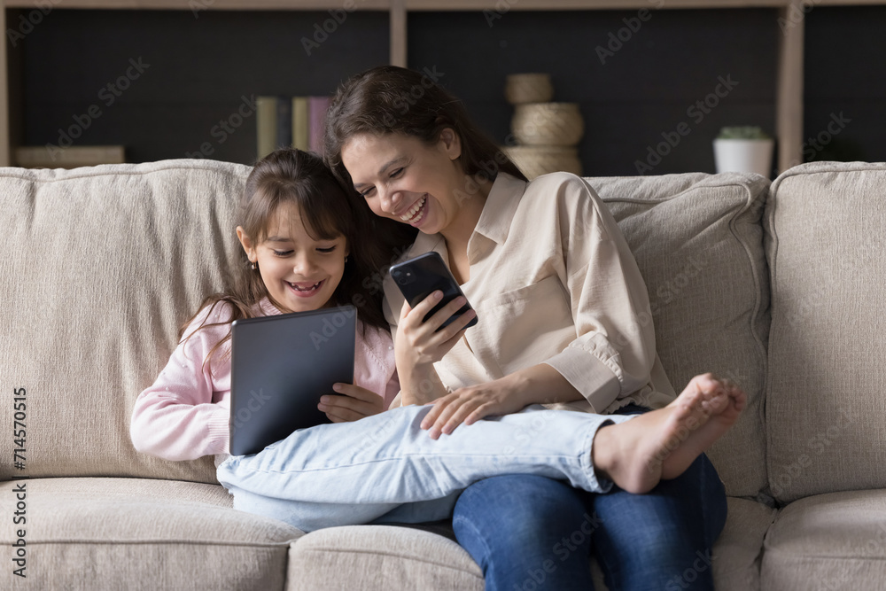 Happy mother and little child using modern technologies seated on sofa, holding digital tablet and smartphone discuss new supplication, having fun, play games laughs on on-line videos. Tech overuse Happy mother and little child using modern technologies seated on sofa, holding digital tablet and smartphone discuss new supplication, having fun, play games laughs on on-line videos. Tech overuse