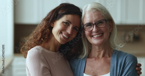 Happy senior mom and adult daughter woman posing for family portrait at home, looking at camera with toothy smiles, laughing, hugging, standing close with faces touch, enjoying warm relationship photo