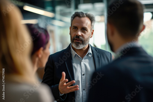 Bearded businessman in formal suit gesticulating and talking with focused peoples while standing in office during work time photo