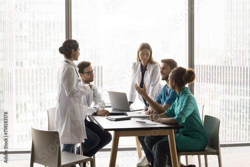 Young clinic chief man talking to diverse staff of doctors at meeting table, giving consultation of expert to listening multiethnic practitioners. Medical colleagues discussing job, treatment case