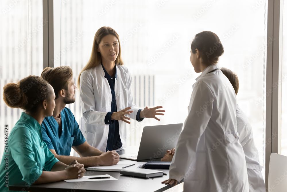 © fizkes - Two positive diverse female doctor colleagues in whites coats discussing healthcare problems, medical job case on professional council, standing at team meeting table, talking