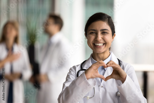 Cheerful young Indian doctor woman making hand heart, looking at camera with happy toothy smile, showing romantic gesture of love, care, health protection, cardiology healthcare photo