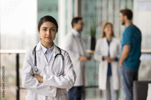 Serious confident young Indian doctor woman posing for professional portrait with arms crossed, looking at camera, standing in clinic hall space with medical team in blurred background
