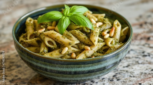  a bowl of pasta with pesto, pine nuts, and basil on top of a granite countertop with a green leaf on top of the pasta in the bowl.